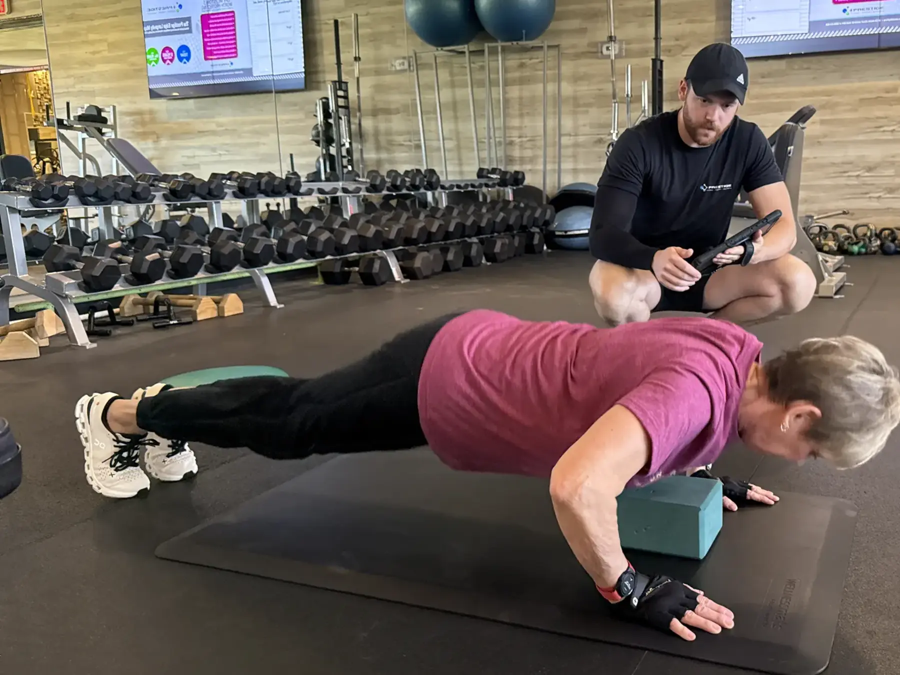 A Prestige Fitness coach guiding a client while the Prestige Edge plan is visible in the studio